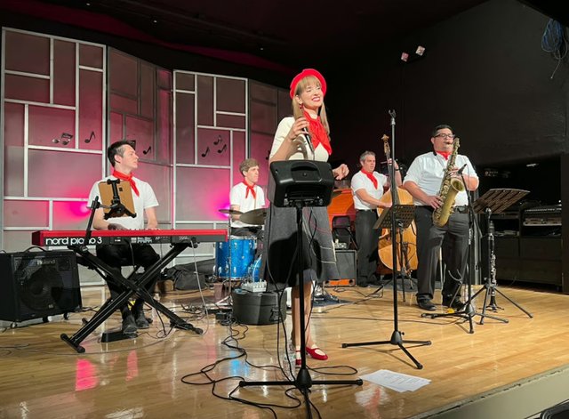Ashley Anne in a red beret and neckerchief singing with The Carnations in white shirts and red scarves, Parisian café style
