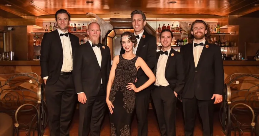Ashley Anne in a beaded flapper gown and feathered headpiece with The Carnations in black tie at an ornate vintage bar