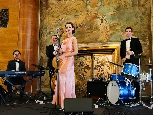 Ashley Anne in a blush 1930s gown performing with upright bass and drums beside the ornate Art Deco fireplace of the Queen Mary's Grand Salon